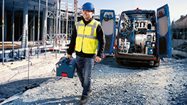 A construction worker in a yellow safety vest and blue hard hat is carrying a toolbox and walking towards a blue van.