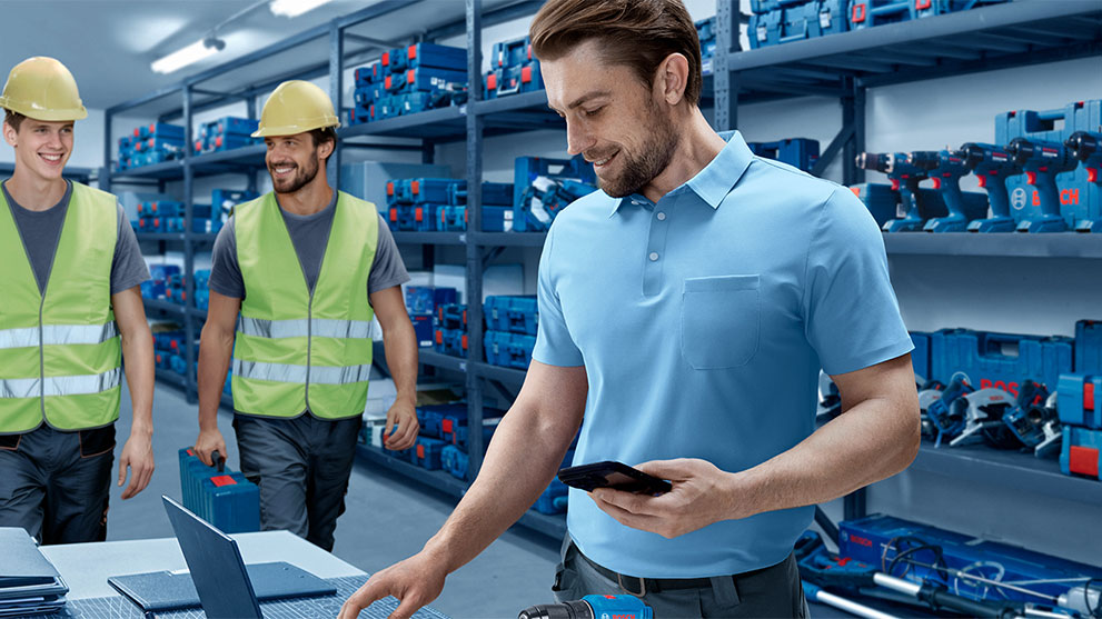 Three men in work clothes stand in a warehouse full of blue toolboxes. The man on the right, wearing a light blue polo, is looking at his phone. The others are wearing yellow helmets and reflective vests. The mood is friendly and cooperative.