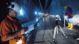 Two construction workers in a tunnel, one wearing a helmet, the other with a measuring device.
