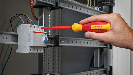 A hand holds a red Bosch Professional screwdriver next to a switch box.