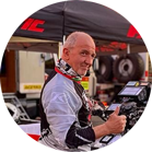 A man in a helmet and racing suit gives a thumbs up in front of a tent and tools.