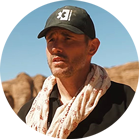 A man with a black hat and scarf stands in front of a rocky landscape.