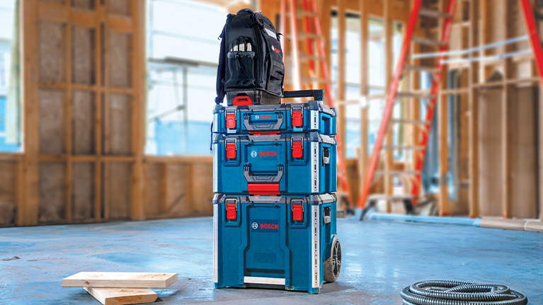 Three Bosch toolboxes in blue and black, with a backpack on top, in a construction environment.