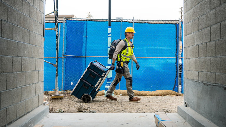 A construction worker wearing a helmet and safety vest is pulling a wheeled toolbox through a construction site.