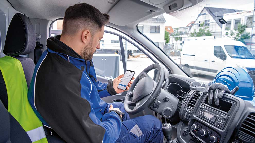A man in a blue work suit sits in a van and looks at his smartphone.